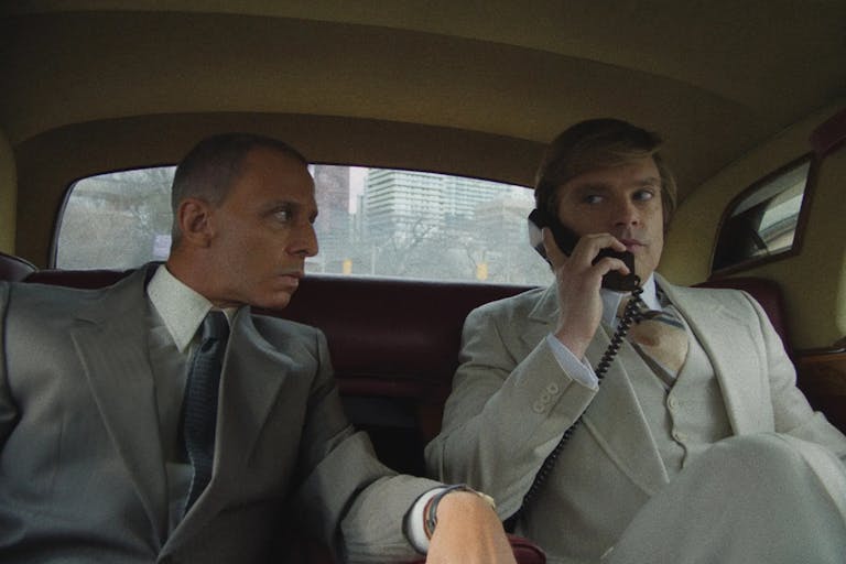Two white men in expensive looking suits sitting in the back of a car