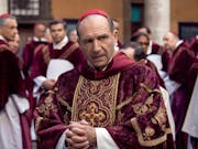 A middle aged white man in red velvet cardinal clothes surrounded by other men in similar clothing