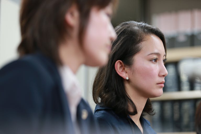 A young Japanese woman in a suit, sitting in a lawyers office beside another Japanese woman blurred in the foreground, with a troubled face