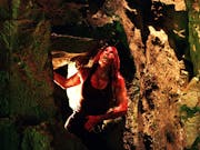 A white woman covered in blood, crouched down in a claustrophobic cave looking up
