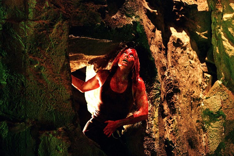 A white woman covered in blood, crouched down in a claustrophobic cave looking up