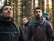 A group of one Asian man and three white men in outdoor hiking clothing standing in a forest looking unnerved