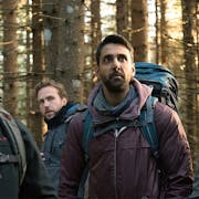 A group of one Asian man and three white men in outdoor hiking clothing standing in a forest looking unnerved