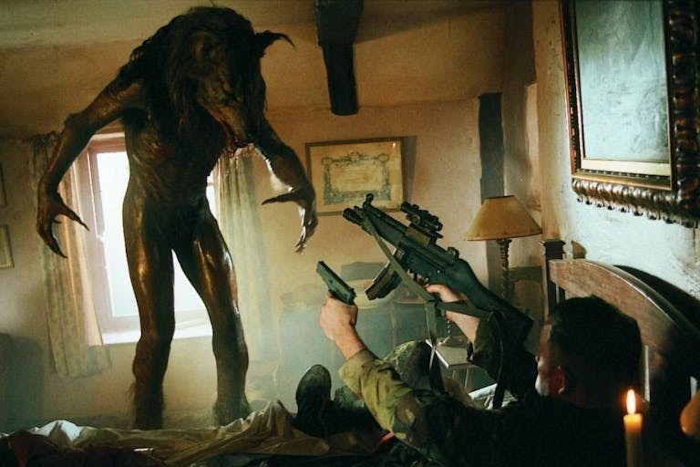A werewolf towers over a man in uniform is on his back pointing two guns at it while in a small cabin