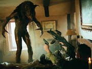 A werewolf towers over a man in uniform is on his back pointing two guns at it while in a small cabin