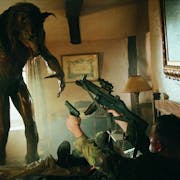 A werewolf towers over a man in uniform is on his back pointing two guns at it while in a small cabin