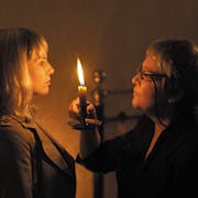 An middle aged white woman holds up a candle to another white woman as they face each other in the dark