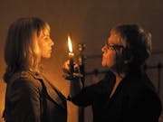 An middle aged white woman holds up a candle to another white woman as they face each other in the dark