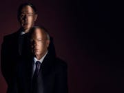 Two white men in suits staring straight ahead in the shadows with a single strip of light across their face