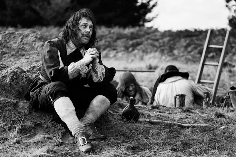 Black and white still of a white man in 17th Century English quaker style clothing sitting in a field with his hands crossed pleading as two man are behind him digging a hole