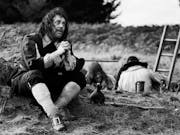 Black and white still of a white man in 17th Century English quaker style clothing sitting in a field with his hands crossed pleading as two man are behind him digging a hole