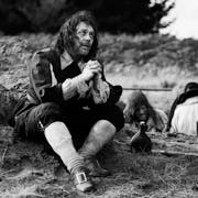 Black and white still of a white man in 17th Century English quaker style clothing sitting in a field with his hands crossed pleading as two man are behind him digging a hole