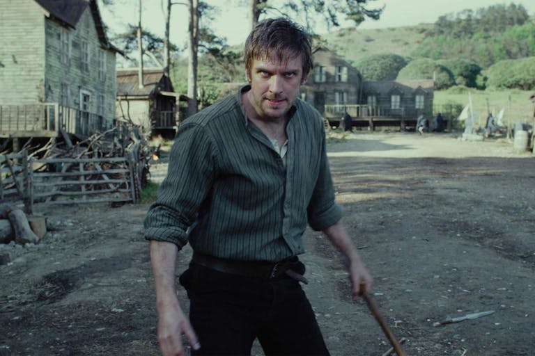 A white man in a small, rural, period village, standing off looking manic holding a large stick