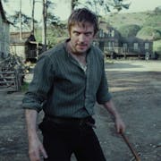 A white man in a small, rural, period village, standing off looking manic holding a large stick