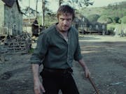 A white man in a small, rural, period village, standing off looking manic holding a large stick