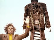 A white man with wild hair wearing a tweed blazer and a yellow jumper, arms outstretched in front a large wooden effigy of a man