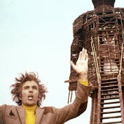 A white man with wild hair wearing a tweed blazer and a yellow jumper, arms outstretched in front a large wooden effigy of a man