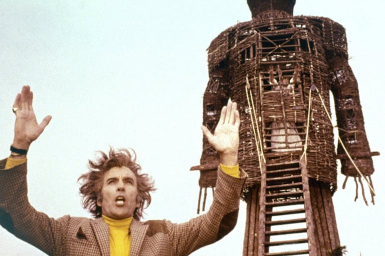 A white man with wild hair wearing a tweed blazer and a yellow jumper, arms outstretched in front a large wooden effigy of a man
