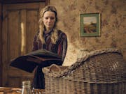 A white woman in a dated country house holding a large book looking tired and haunted at a wicker baby basket