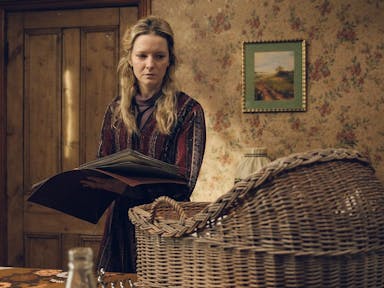 A white woman in a dated country house holding a large book looking tired and haunted at a wicker baby basket