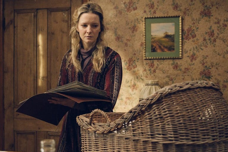 A white woman in a dated country house holding a large book looking tired and haunted at a wicker baby basket