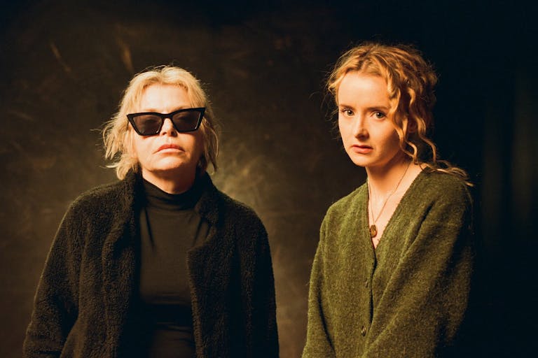 Two women face the camera. One has blond hair, dark sunglasses and is wearing black. The second woman is wearing green and has curly light red hair.