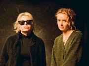 Two women face the camera. One has blond hair, dark sunglasses and is wearing black. The second woman is wearing green and has curly light red hair.