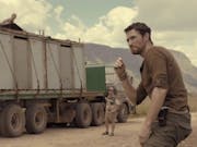 A white man in casual army combat clothes looking suspiciously over to the side with a large covered truck behind him