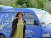A middle aged brown woman in a light green salwar kameez top and long black cardigan standing in front of a tiny light blue catering van