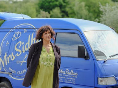 A middle aged brown woman in a light green salwar kameez top and long black cardigan standing in front of a tiny light blue catering van 