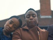 Close up a young Black girl with her head on a Black woman's shoulder, standing next to a tall young Black man in a suit, all looking down looking devastated