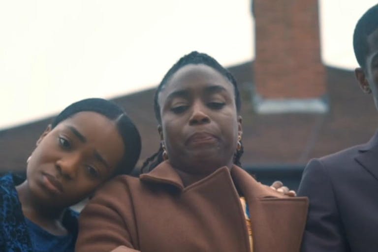 Close up a young Black girl with her head on a Black woman's shoulder, standing next to a tall young Black man in a suit, all looking down looking devastated