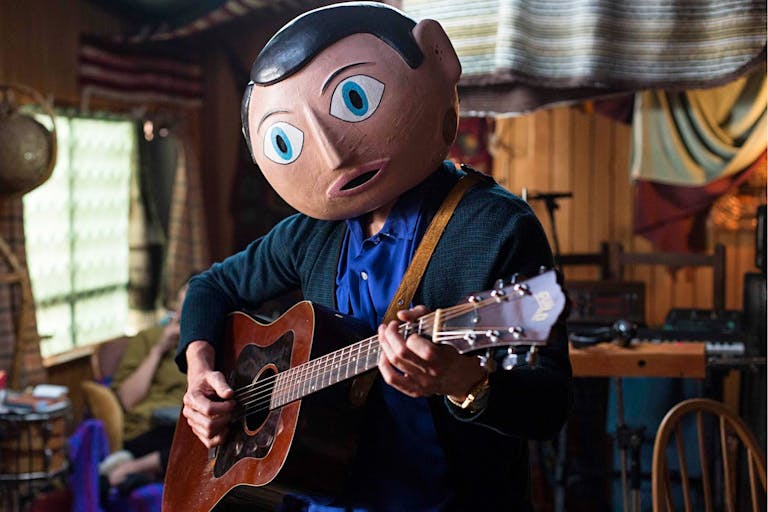 A white man in a large paper mache head with large blue eyes, playing a guitar in a cluttered house