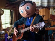 A white man in a large paper mache head with large blue eyes, playing a guitar in a cluttered house
