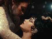 A white man with monster-like features, beard and long hair dancing with a woman in sequin white dress and black hair with a white streak in a high updo
