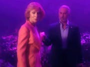 Dreamy purple and pink lighting over a white woman in a pink suit and a white man in a dark suit
