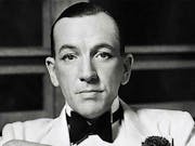 Black and white still of a slim faced white man with slicked back hair, white tux and black bowtie