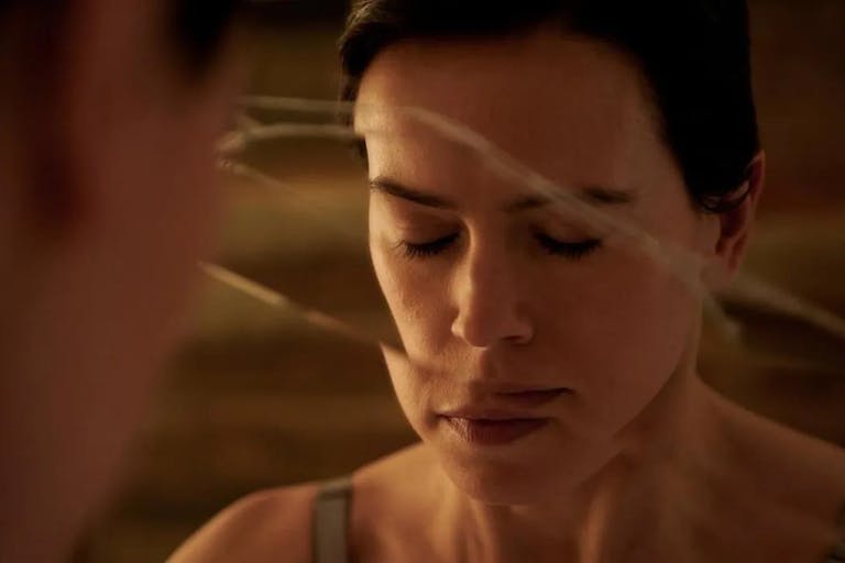 A white woman with cropped brown hair with closed eyes facing a fractured mirror