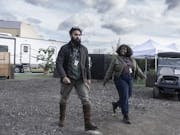 A tall bearded brown man in wellies and a short Black woman wearing lanyards walking through a film set with trailer and buggy in the background