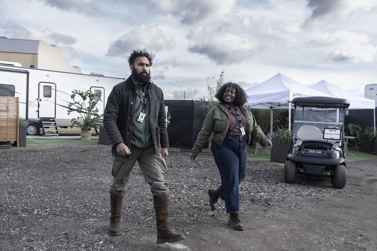 A tall bearded brown man in wellies and a short Black woman wearing lanyards walking through a film set with trailer and buggy in the background