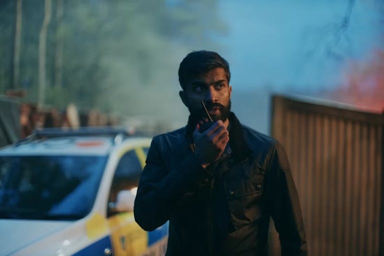 A brown, bearded man detective talking into a radio device with a police car in the background