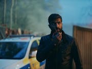 A brown, bearded man detective talking into a radio device with a police car in the background