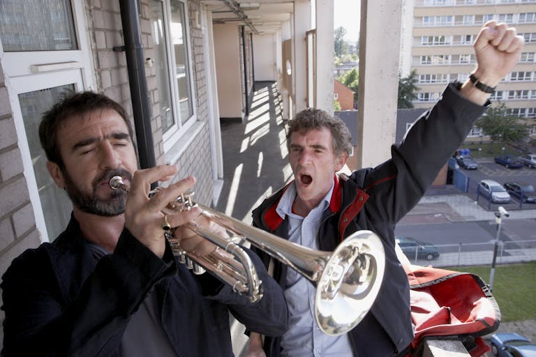 A white man playing the trumpet outside a second floor flat as a white man stands behind triumphantly with his arm in the air