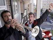 A white man playing the trumpet outside a second floor flat as a white man stands behind triumphantly with his arm in the air
