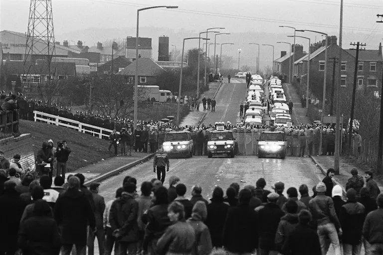 Black and white still of a long street in small rural town with a group of people facing off a wall of police and police cars