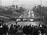 Black and white still of a long street in small rural town with a group of people facing off a wall of police and police cars