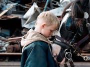 A young white boy stands in a scrap yard with his head rested against a workhorses head