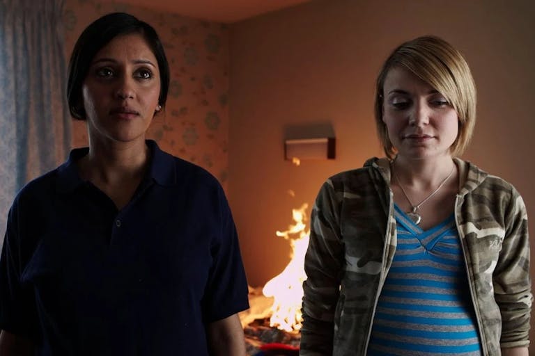 A young British Asian woman and a young white pregnant woman stand in a small room looking to camera as a small fire burns in the background
