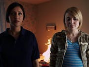 A young British Asian woman and a young white pregnant woman stand in a small room looking to camera as a small fire burns in the background