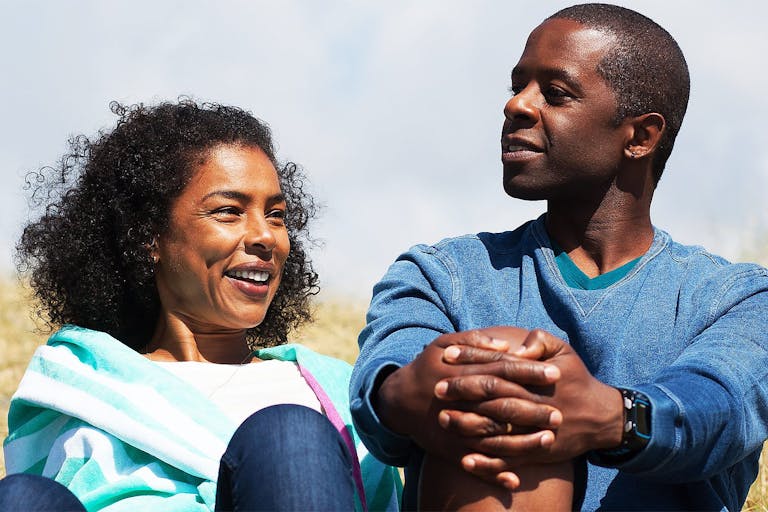 A Black woman and Black man sit on a beach smiling